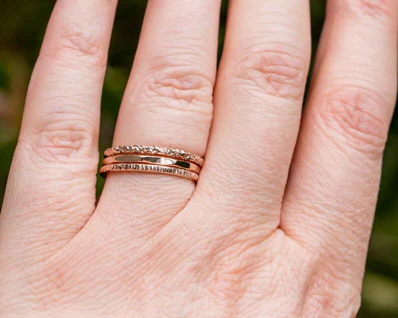 May include: Three gold-toned stacking rings on a finger. The rings have different textures, including a hammered finish and a textured band.