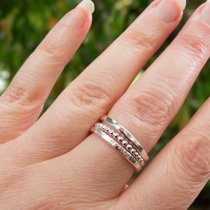 Puede incluir: Tres anillos de plata apilables en un dedo. Los anillos son de diferentes estilos, uno es una banda simple, otro tiene una textura de cuentas y el otro tiene una textura martillada.