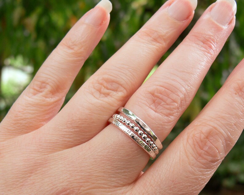 Puede incluir: Tres anillos de plata apilables en un dedo. Los anillos son de diferentes estilos, uno es una banda simple, otro tiene una textura de cuentas y el otro tiene una textura martillada.