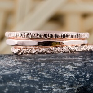 May include: Three rose gold stacking rings with textured surfaces. The rings are stacked on top of each other and are sitting on a gray rock.