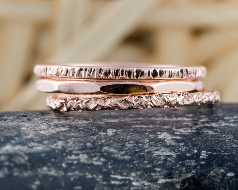 May include: Three rose gold stacking rings with textured surfaces. The rings are stacked on top of each other and are sitting on a gray rock.