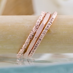 May include: Three rose gold stacking rings with a textured finish. The rings are stacked on a light brown surface.