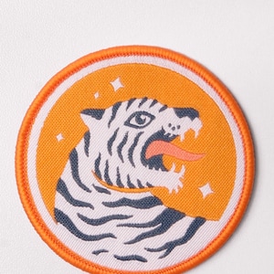 Può includere: Toppa ricamata arancione e bianca con un design a testa di tigre. La tigre ha un viso bianco con strisce nere e una lingua arancione.