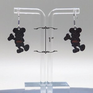 May include: A pair of black Mickey Mouse silhouette earrings. Each earring is approximately 1 inch in length. The earrings hang from silver-colored fish hook ear wires. The earrings are displayed on a clear acrylic earring stand.