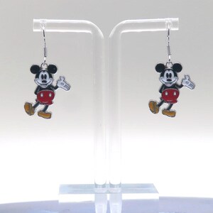 May include: A pair of Mickey Mouse earrings. Each earring features a black and white cartoon character with a red shorts, yellow shoes, and white gloves. The earrings are suspended from silver-colored fish hook ear wires.