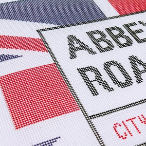May include: A white canvas needlepoint with a black and white sign that reads "Abbey Road" and "City of" in red. The sign is bordered by a black frame. The background is a red, white, and blue design.