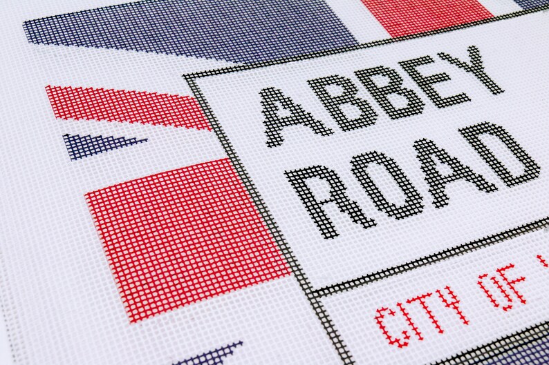 May include: A white canvas needlepoint with a black and white sign that reads "Abbey Road" and "City of" in red. The sign is bordered by a black frame. The background is a red, white, and blue design.