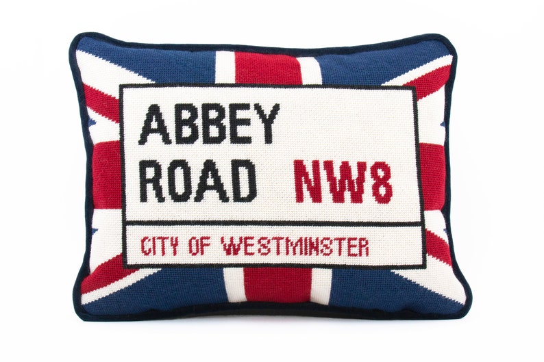 May include: A decorative pillow with a red, white, and blue Union Jack design. The pillow features a white rectangular patch with black text that reads "Abbey Road NW8 City of Westminster".