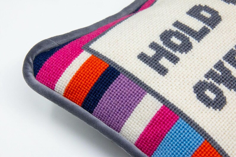 May include: A white pillow with a gray border and colorful stripes on the bottom. The pillow is embroidered with the text "Hold on" in gray.