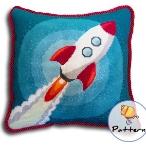 May include: A blue and white needlepoint pillow with a red and white rocket ship design. The rocket ship is flying through a starry sky with a trail of smoke behind it.
