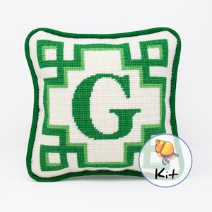 May include: Green needlepoint pillow with a white background and a large green letter G in the center. The pillow has a green border and a small image of a hermit crab in the lower right corner with the word "Kit" written below it.