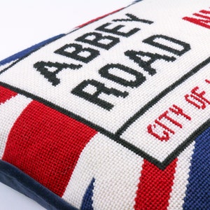 May include: A white pillow with a blue, red, and white Union Jack pattern. The pillow has a black embroidered sign that reads "Abbey Road" and "City of Westminster".