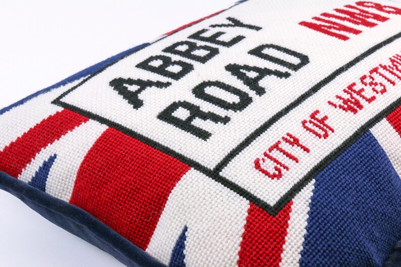 May include: A white pillow with a blue, red, and white Union Jack pattern. The pillow has a black embroidered sign that reads "Abbey Road" and "City of Westminster".