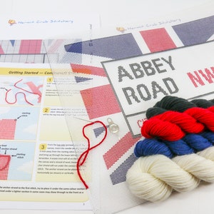 May include: A cross-stitch kit featuring the iconic Abbey Road street sign with the Union Jack flag in the background. The kit includes a white canvas, embroidery floss in red, white, blue, and green, a needle, and instructions.