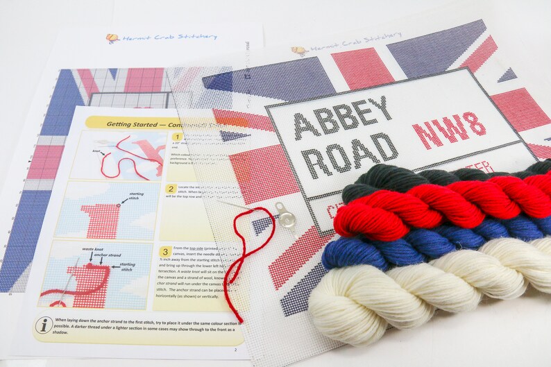 May include: A cross-stitch kit featuring the iconic Abbey Road street sign with the Union Jack flag in the background. The kit includes a white canvas, embroidery floss in red, white, blue, and green, a needle, and instructions.