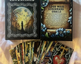 May include: A tarot card deck and two boxes are displayed. The boxes are black with white text that reads "GOOD MOJO LENORMAND". The deck is fanned out, revealing colorful illustrations. The text "GOOD MOJO LENORMAND ORACLE" is on one box.
