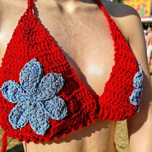 May include: A bright red crochet halter top featuring a large, light blue six-petaled flower appliqué on the left cup and a smaller matching flower on the right cup. The garment is constructed with a textured yarn stitch and includes thin straps.