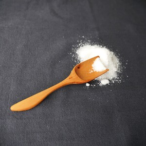Könnte beinhalten: Ein Holzlöffel, gefüllt mit weißen Granulaten, liegt neben einem kleinen Haufen derselben Substanz. Der Löffel ist hellbraun und hat einen gebogenen Griff. Der Hintergrund ist ein dunkelgrauer Stoff.