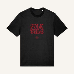 Pode incluir: Uma t-shirt preta de manga curta com o texto vermelho "FOLK TALE" impresso numa fonte estilizada no peito. Abaixo do texto principal, um pequeno gráfico vermelho em forma de diamante contém o texto "FOLK TALE" repetido em tamanho muito pequeno.