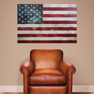 May include: A distressed American flag wall decal with red, white, and blue colors. The flag is hanging on a beige wall above a brown leather armchair.