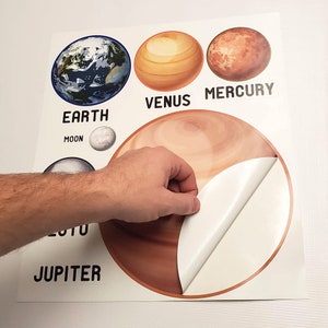 May include: A set of colorful planet stickers with the names "Earth", "Moon", "Venus", "Mercury", and "Jupiter". The stickers are on a white background and one of the stickers is being peeled off.