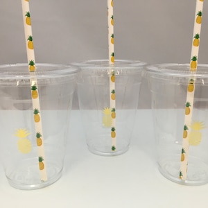 May include: Three clear plastic cups with pineapple patterned straws. The cups are empty and have a white background.