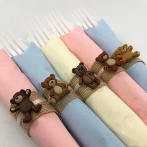 May include: Pastel-coloured napkins, each rolled and secured with a teddy bear button and a twine and ribbon band. Clear plastic forks are inserted into the napkins, suitable for a party or event.