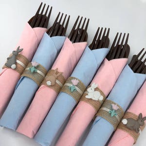 May include: A set of six pink and blue napkins with brown forks wrapped inside. Each napkin has a burlap napkin ring with a small bunny charm attached.