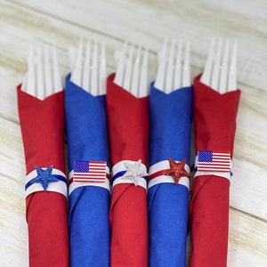 May include: Six red, white, and blue napkins with forks wrapped inside. Each napkin has a white ribbon with a red, white, and blue star and a small American flag.