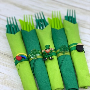 May include: Five green plastic forks wrapped in green napkins with festive St. Patrick's Day themed decorations. The decorations include a rainbow, shamrock, leprechaun, and pot of gold.