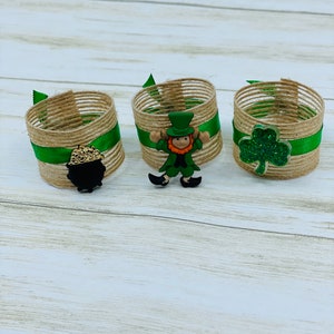 May include: Three brown and green burlap napkin rings with green ribbon and a different St. Patrick's Day themed embellishment on each. The embellishments are a leprechaun, a shamrock, and a pot of gold.