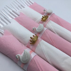 May include: Pink and white napkins with white swan napkin rings with gold crowns. The napkin rings are tied with a pink ribbon.