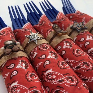 May include: Red and white bandana wrapped around blue plastic forks with cowboy-themed embellishments, including a sheriff's star with "The Law" written on it, a cowboy boot, and a star.