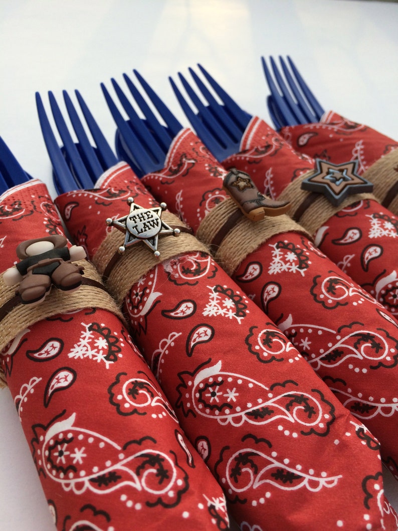 May include: Red and white bandana wrapped around blue plastic forks with cowboy-themed embellishments, including a sheriff's star with "The Law" written on it, a cowboy boot, and a star.