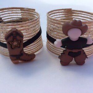 May include: Two brown burlap napkin rings with black ribbon accents. One ring has a brown cowboy boot with a star on it. The other ring has a brown cowboy figure with a brown hat and black shirt.
