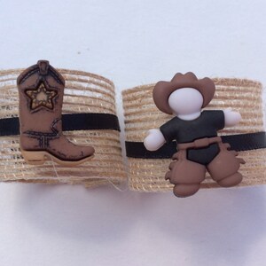 May include: Two brown burlap napkin rings with black ribbon accents. One ring features a brown cowboy boot with a gold star, the other ring features a brown cowboy figurine with a black hat and brown chaps.