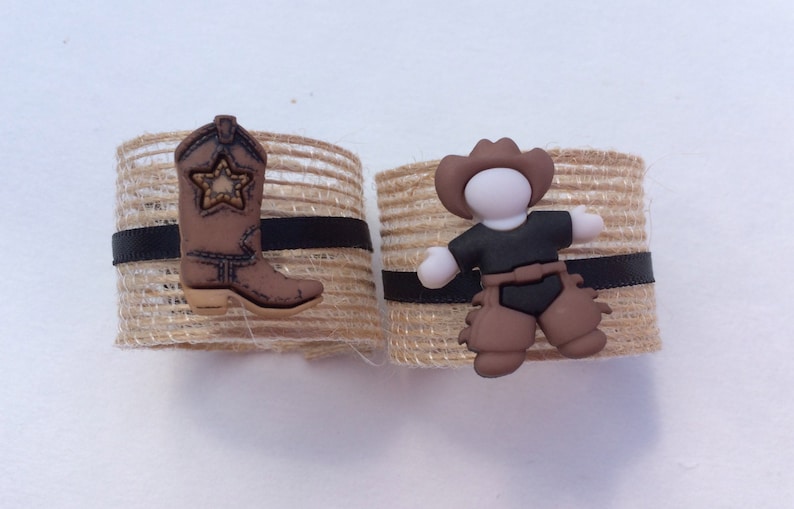 May include: Two brown burlap napkin rings with black ribbon accents. One ring features a brown cowboy boot with a gold star, the other ring features a brown cowboy figurine with a black hat and brown chaps.