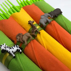 May include: Four green, orange, and yellow napkins with animal-shaped napkin rings. The rings are a zebra, lion, giraffe, and elephant.