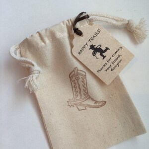 May include: A small, beige drawstring bag with a brown ribbon and a brown paper tag. The tag has the text "Happy Trails!" and a silhouette of a cowboy roping a calf. The tag also has the text "Thanks for coming, your friend, Grayson". The bag has a brown cowboy boot design printed on it.
