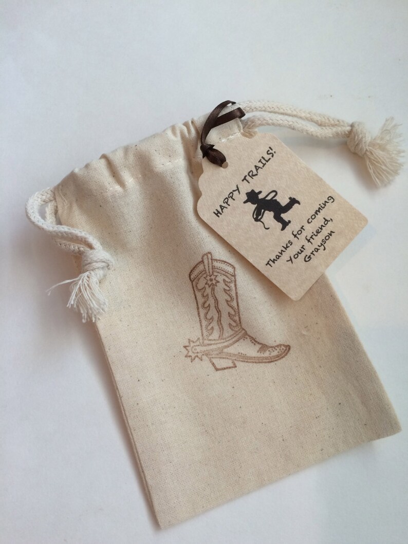 May include: A small, beige drawstring bag with a brown ribbon and a brown paper tag. The tag has the text "Happy Trails!" and a silhouette of a cowboy roping a calf. The tag also has the text "Thanks for coming, your friend, Grayson". The bag has a brown cowboy boot design printed on it.