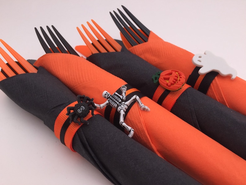 May include: Four orange and black napkins with Halloween-themed napkin rings. The rings feature a spider, a skeleton, a pumpkin, and a ghost.