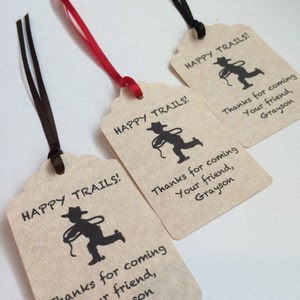 May include: Three brown paper gift tags with black ribbon. Each tag has a silhouette of a cowboy with a lasso and the text "HAPPY TRAILS!" and "Thanks for coming, Your friend, Grayson".