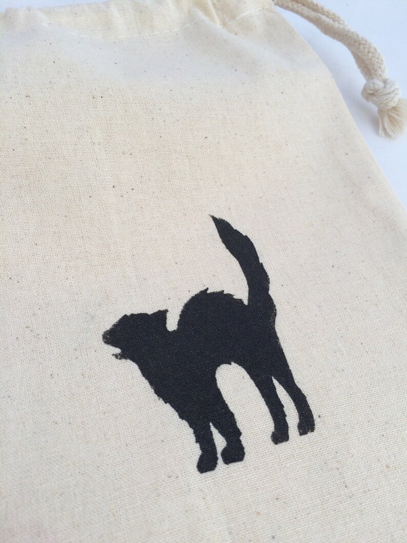 K&ouml;nnte beinhalten: Eine wei&szlig;e Baumwolltasche mit Kordelzug, auf der eine schwarze Silhouette einer Katze aufgedruckt ist.