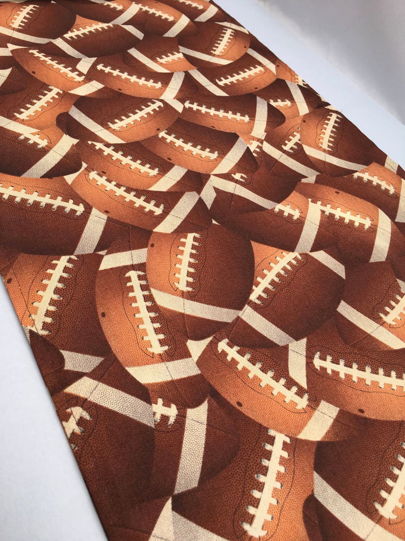 May include: A brown and cream fabric with a repeating pattern of brown footballs. The footballs have white laces and a textured surface.