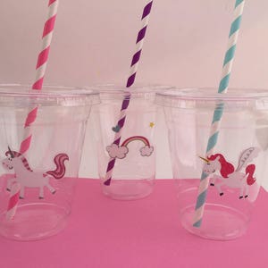 May include: Three clear plastic cups with unicorn and rainbow designs. Each cup has a colourful striped straw.