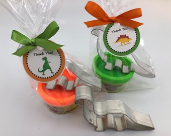 Puede incluir: Dos bolsas de plástico transparentes con cintas verdes y naranjas atadas alrededor. Cada bolsa contiene un pequeño recipiente de Play-Doh y un cortador de galletas con forma de dinosaurio. Las bolsas tienen una etiqueta redonda adjunta con el texto "¡Gracias!" y una imagen de un dinosaurio.