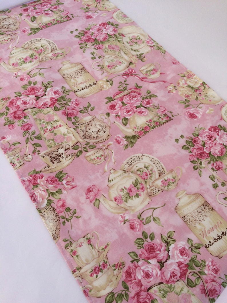 May include: Pink table runner with a floral pattern featuring roses and teacups. The pattern includes the words "Roses" and "Flowers".