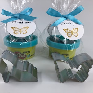 May include: Two small plastic containers of Play-Doh, each with a blue lid and a butterfly-shaped cookie cutter. The containers are packaged in clear cellophane bags with blue ribbons and a thank you tag with a gold butterfly design.
