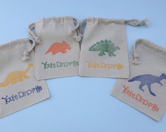 Puede incluir: Cuatro pequeñas bolsas de tela beige con cordón, con siluetas de dinosaurios de diferentes colores y el texto "You're Dino-mite!" impreso en cada una.