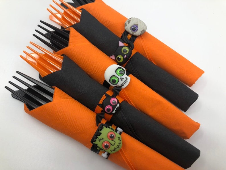 May include: Set of five orange and black Halloween napkin rings with different spooky character designs. Each ring features a different character, including a mummy, a cat, a skull, a monster, and a Frankenstein.